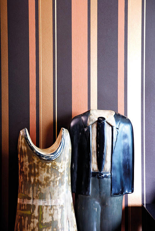 A pair of “headless” dress and tuxedo sculptures by Russian artist Yuri Zatarain stand in front of an accent wall with tan and gold striped wallpaper in the living room.