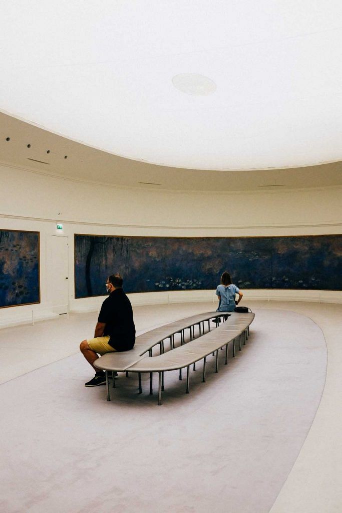 Two persons sitting on a round bench in a minimalist art gallery. Photo by Pexels Adrien Olichon.